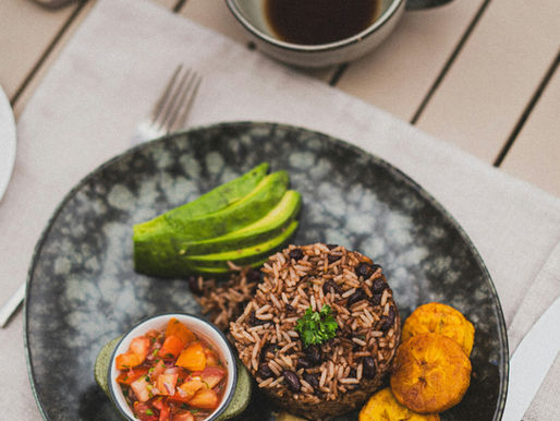 Plate with rice, beans, avocado, plantains, salsa, and tortillas. Coffee cup and water glass on a wooden table. Calm outdoor setting.