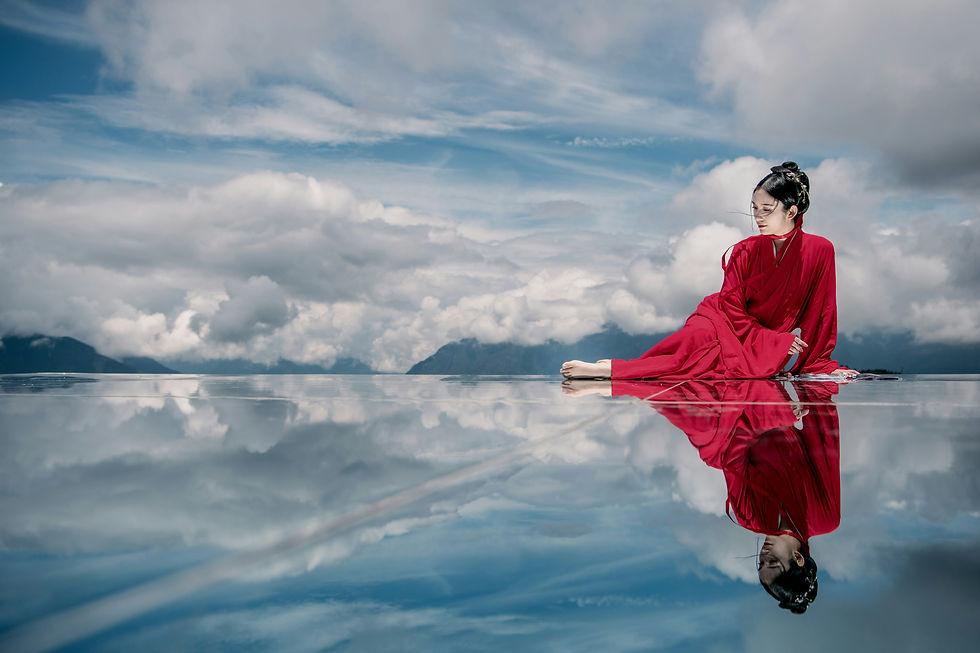 Woman in a flowing red dress seated beside reflective water beneath an expansive sky, symbolizing personal reflection and clarity.