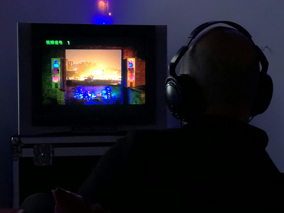 Person with headphones watches a TV screen displaying a cityscape with vibrant lights in a dim room. Text in a foreign script appears.