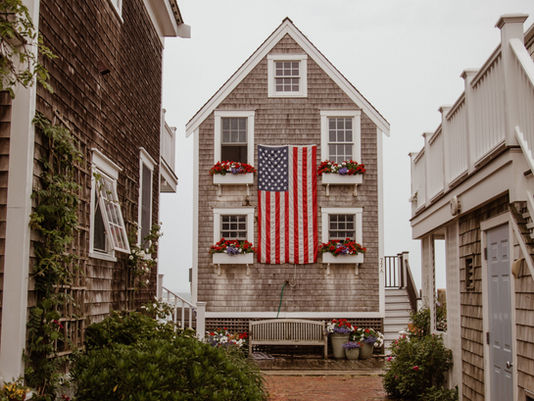 A charming coastal home adorned with blooming flowers and an American flag stands prominently along a quaint brick pathway, capturing the essence of small-town Americana.