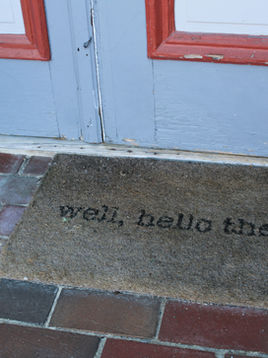 A doormat with "well, hello there" text lies on a red brick porch, in front of a blue door with red trim, creating a welcoming vibe.