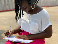 Femme assise, écouteurs noirs, écrit dans un carnet avec des pantalons roses. Fond flou de végétation. Temps ensoleillé, ambiance studieuse.