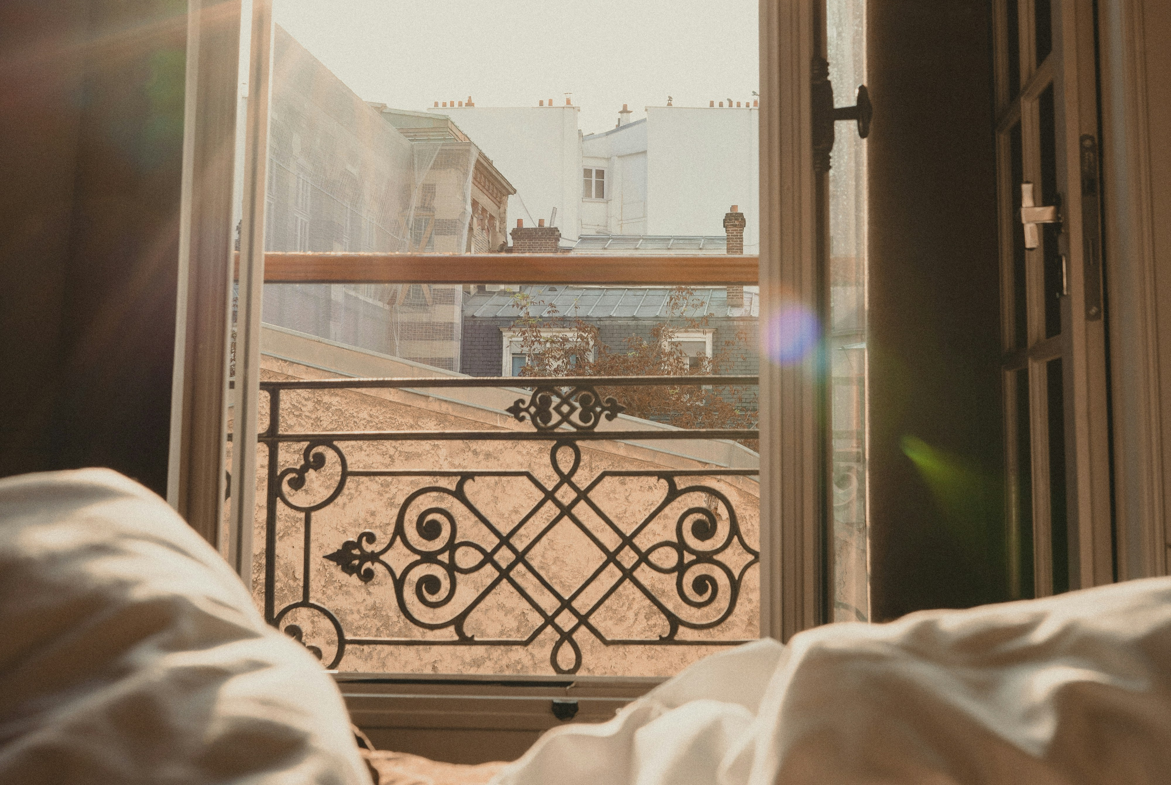 Sunlit bedroom view through open French doors to ornate balcony and rooftops, with white rumpled bedding in foreground.