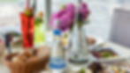 A table with bread, assorted cheeses, and a floral centerpiece overlooks a cityscape. Two colorful drinks in tall glasses add vibrancy.