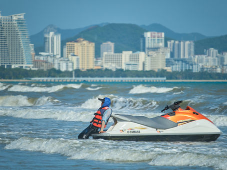 Pessoa com colete salva-vidas e proteção contra o sol empurrando um jet ski Yamaha na beira do mar, com ondas quebrando ao redor e uma cidade com prédios altos e montanhas ao fundo.