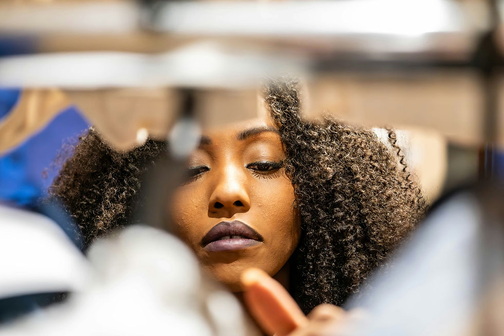 Woman with curly hair and purple lipstick looks down thoughtfully. Blurred background with soft colors suggests an indoor setting.