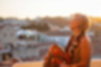 Woman in colorful dress sits on rooftop at sunset, gazing upwards. Cityscape and hot air balloon in the blurred background.