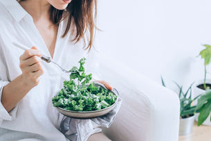 Woman in a white shirt eating kale salad with a fork in a bright room, with potted plants in the background.