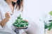Woman in a white shirt eating kale salad with a fork in a bright room, with potted plants in the background.