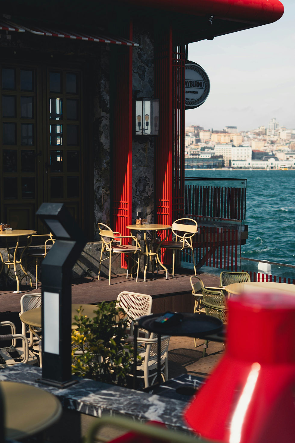 Empty seaside café terrace with beige chairs and red accents, overlooking blue water and a distant city skyline with a round sign