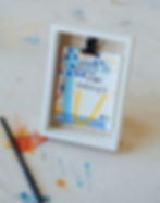Framed sign reading "today is more than enough" with colorful patterns, on a paint-splattered surface beside a black paintbrush.