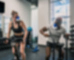 Two people wearing workout masks exercise on bikes in a gym. They're focused and determined. Equipment visible around them.