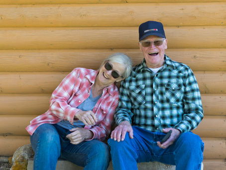 Elderly couple laughing, sitting against a wooden log wall. Woman in pink plaid, man in green plaid with cap. Casual, joyful mood.