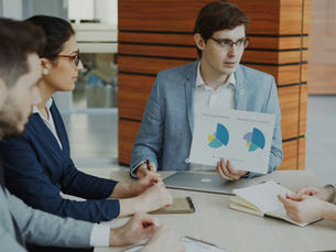 Sales strategy meeting of four people in a meeting room discussing charts. One holds a sales report. Business attire, neutral tones, focused and attentive mood.
