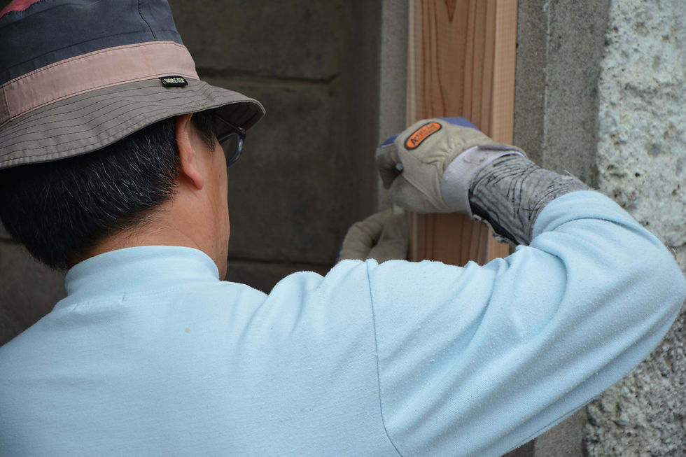 Man in a hat and gloves works on a wooden board against a stone wall. Wearing light blue shirt, focused on task, earthy tones.