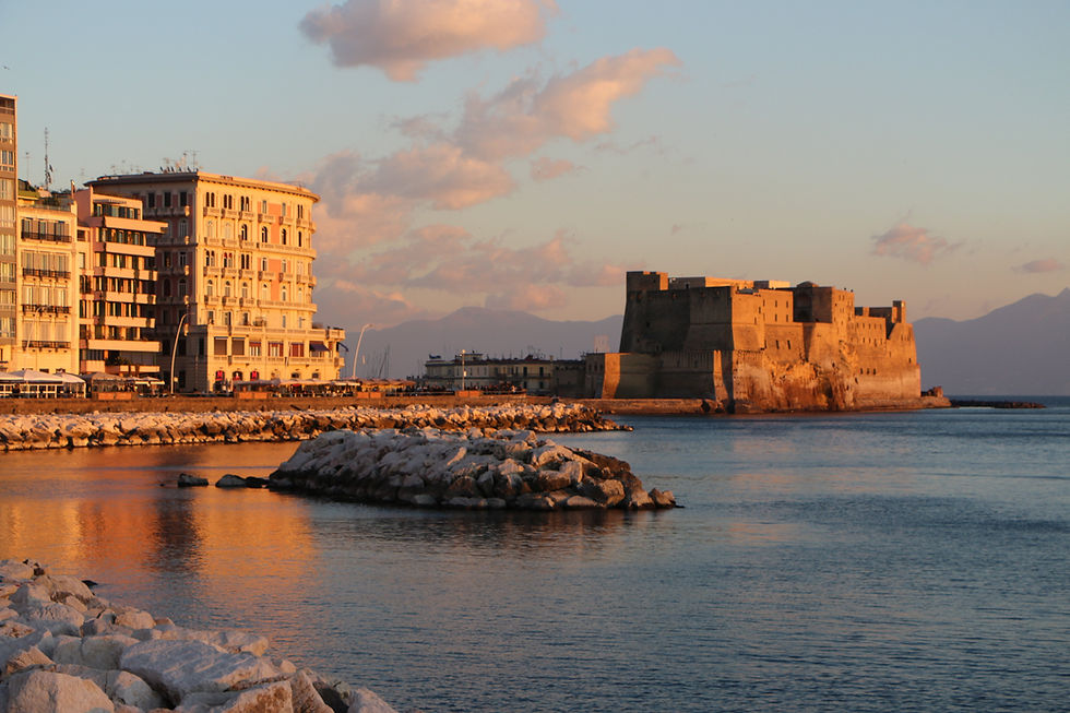 Sunset view of seaside buildings and a historic castle, with a calm sea and clouds painted orange. Peaceful and picturesque setting.