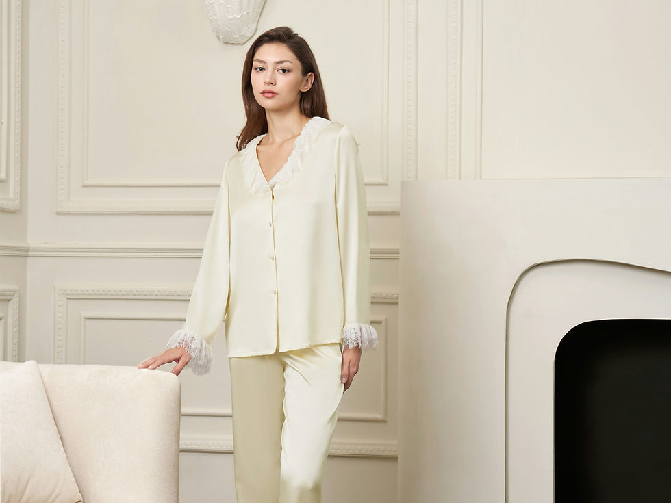 Femme en pyjama crème, debout dans un salon élégant aux murs blancs, regard calme, main sur un fauteuil.