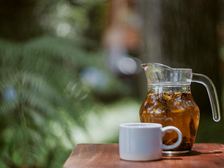 Pitcher of iced tea and white mug on a wooden table. Lush green fern background creates a serene outdoor setting.
