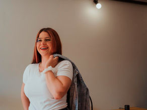 Smiling woman in glasses and white shirt holds a denim jacket over her shoulder in a warm, minimalist room.