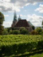Cathedral with tall spires rises above green vineyards and trees under a bright, cloudy blue sky.