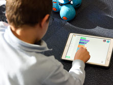 Child using a tablet with colorful programming blocks displayed. Blue robot toy in the background on a dark textured surface.