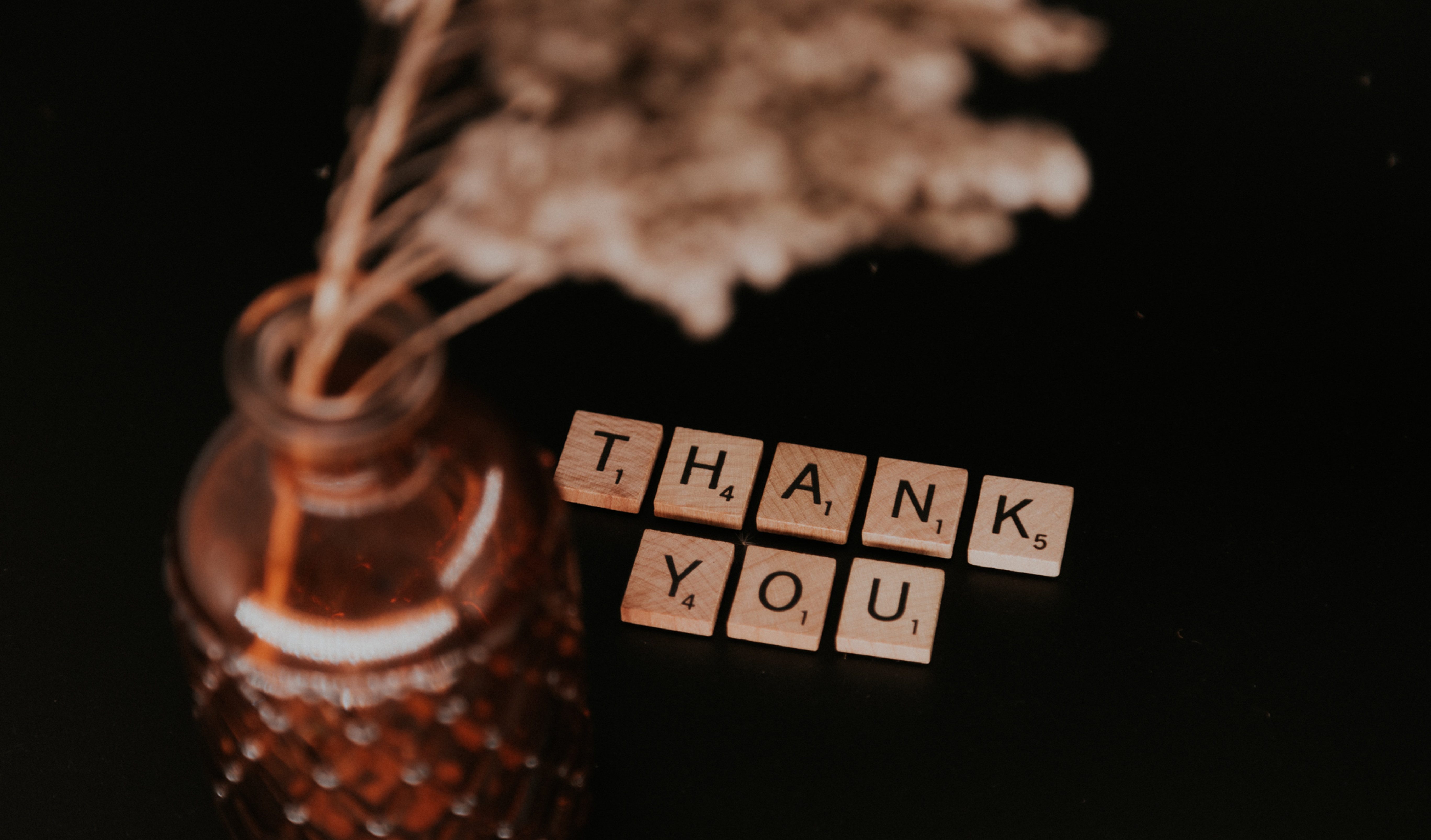 Amber glass bottle with dried flowers beside Scrabble tiles spelling THANK YOU on a dark background. Author Renee Coloman expresses her gratitude for book lovers & readers supporting indie authors. RCwriter-pen-ink-suffering.com