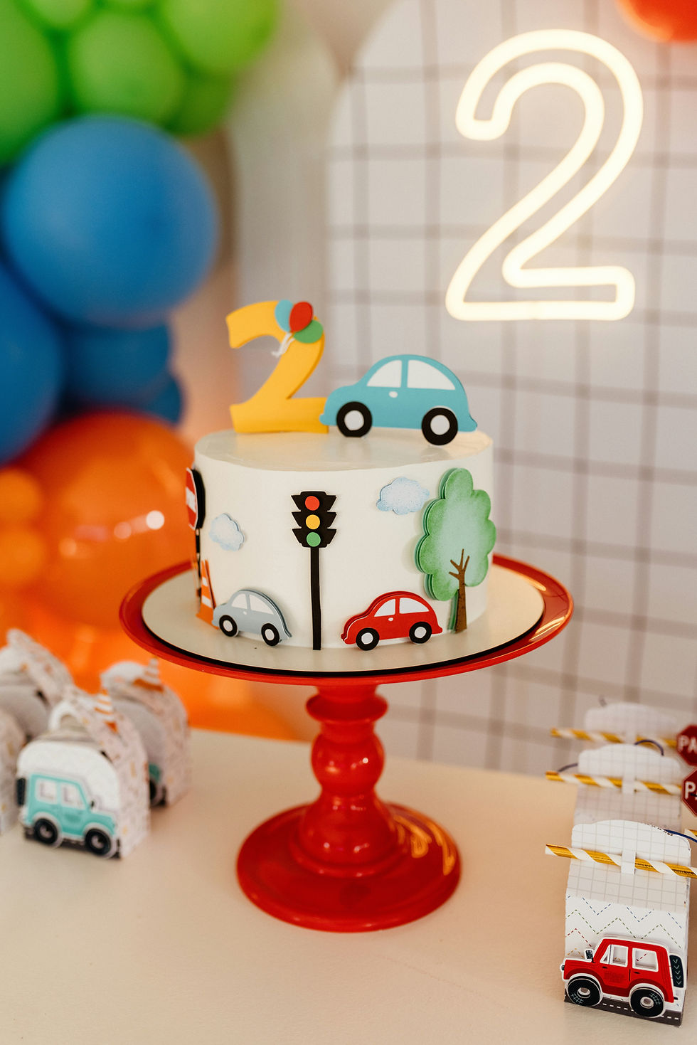 A car cake for a two year old on a red cake stand with balloons in the background