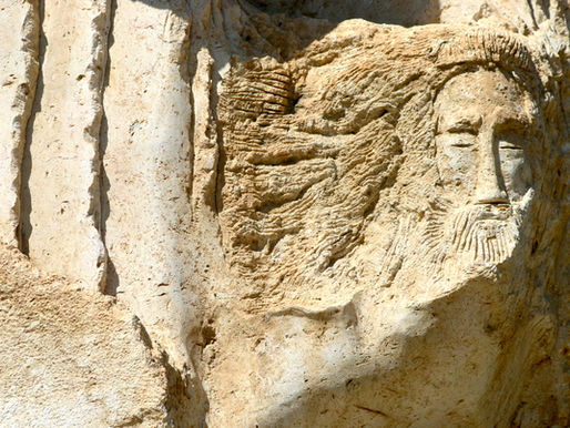 Stone carving of a bearded face with closed eyes on a textured rock surface. Natural light highlights the beige and tan colors. Peaceful mood.