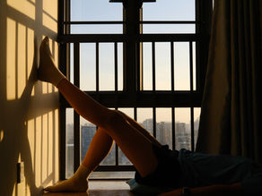 Person lying on the floor with legs against a wall in front of a window. Sunlight casts shadows; the mood is calm and introspective.