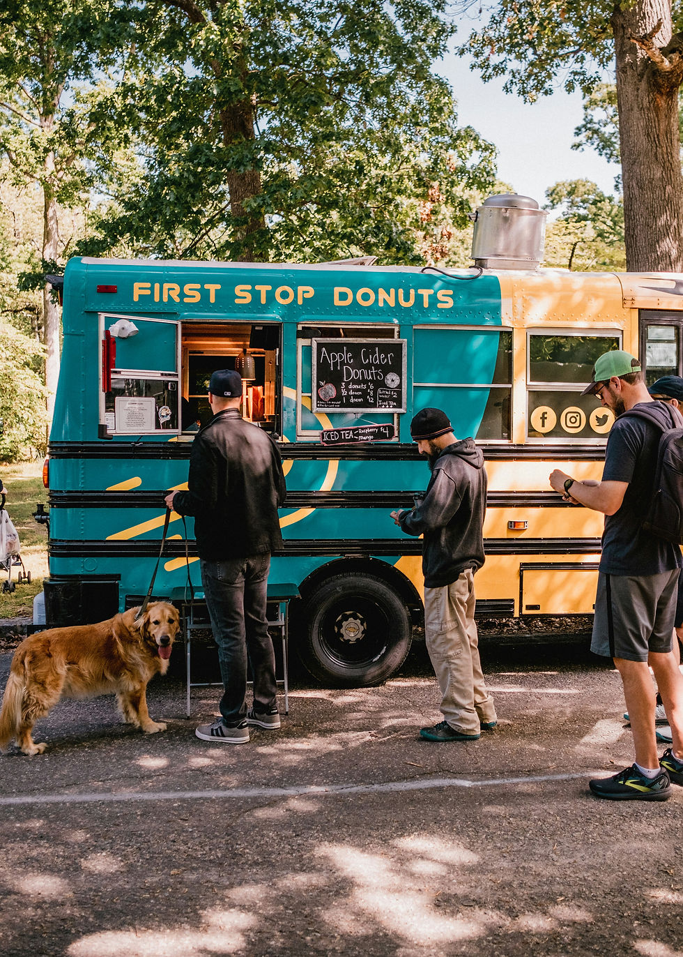 Man with a golden retriever dog orders at a blue and yellow "First Stop Donuts" food truck in a park. Others wait in line, surrounded by trees.