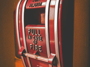 Close-up of a red fire alarm on an orange wall. Text reads "FIRE ALARM" and "PULL IN CASE OF FIRE." Bold colors create urgency.