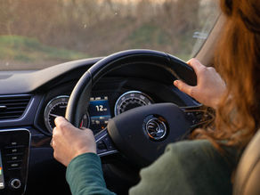 Woman driving a Škoda car at sunset, hands on the wheel, with dashboard text NAV, APP, CAR, MENU and 12 mph visible.
