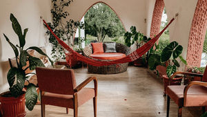 Cozy porch with a red hammock, chairs, plants, and a round wicker seat. Arched windows show lush greenery, evoking a calm, relaxing vibe.