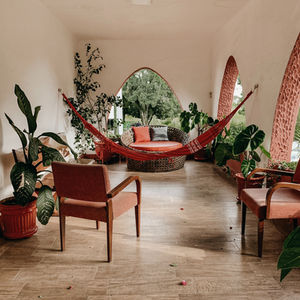 Cozy porch with a red hammock, chairs, plants, and a round wicker seat. Arched windows show lush greenery, evoking a calm, relaxing vibe.