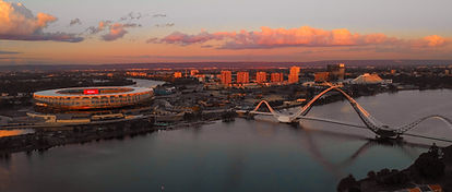 Aerial view of a city at sunset with a large stadium on the left, a modern white arched bridge over water, and buildings in the background under colorful clouds.