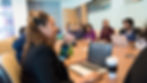 A diverse group in a conference room around a table with laptops. One person raises a hand. Coffee cups and bottles are visible, creating a focused mood.