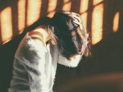 Woman in a soft sweater sits in sunlight with head bowed and hand on hair. Warm, shadowed patterns create a reflective, calm mood.