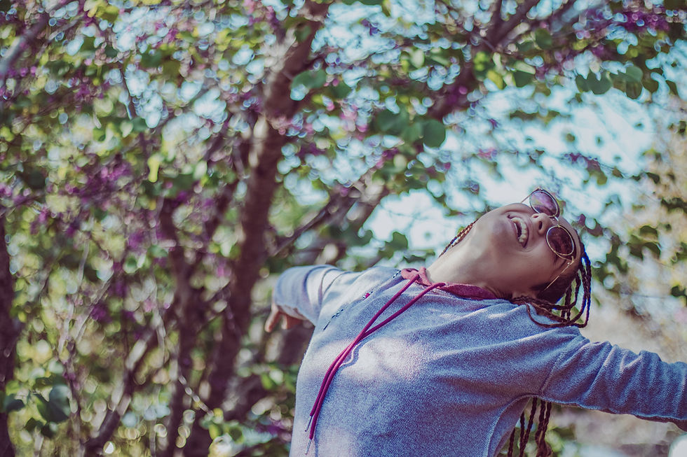 Jeune femme en lunettes de soleil, bras ouverts et souriante, sous des arbres aux feuillages flous et colorés.