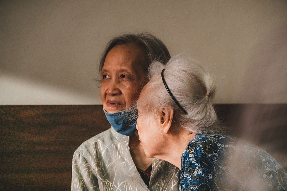 Two elderly women share a tender moment indoors. One whispers intimately. Soft lighting and patterned clothing create a warm atmosphere.
