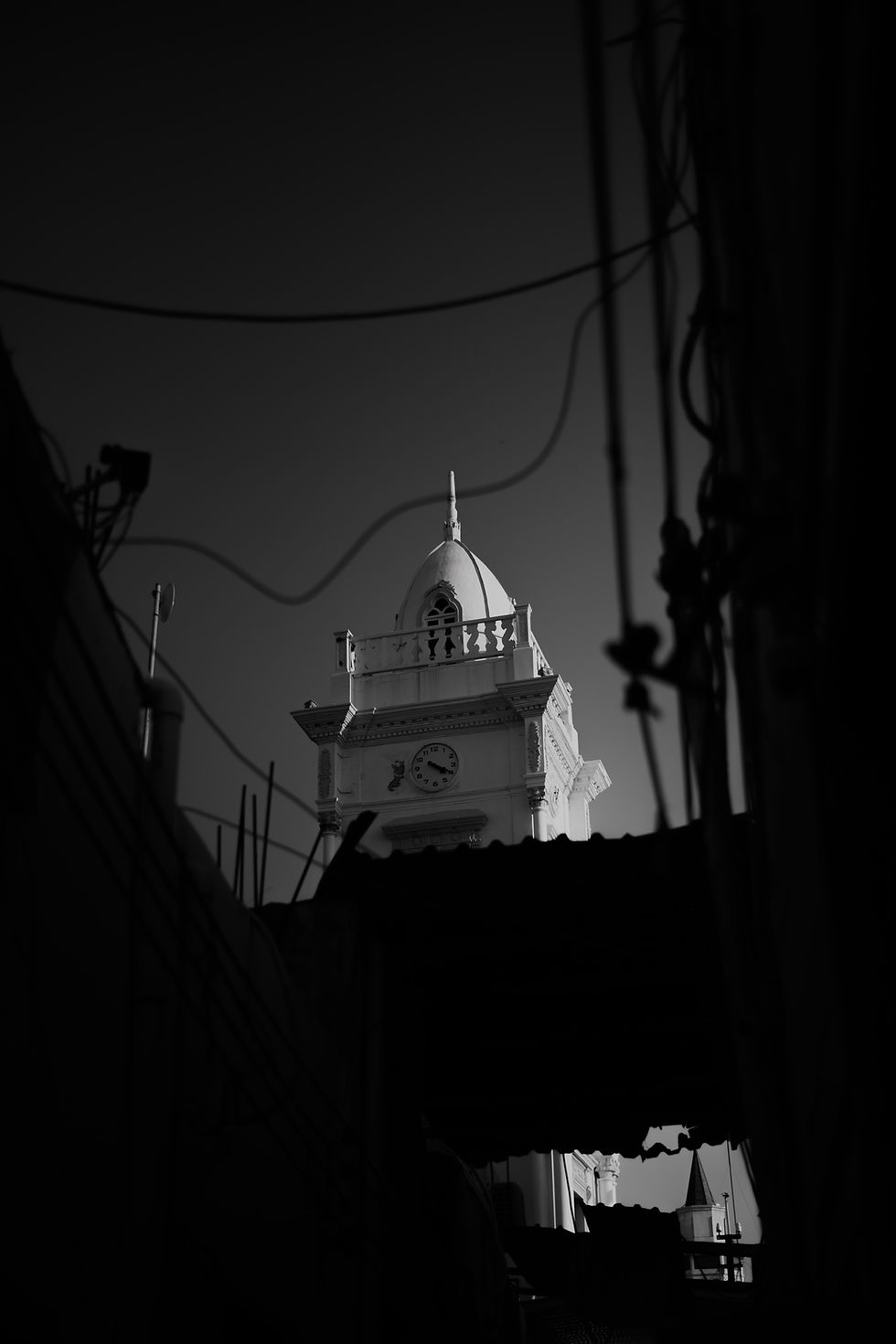 A black and white view of a historic clock tower in Libya, partially obscured by dark silhouettes of surrounding structures and power lines.