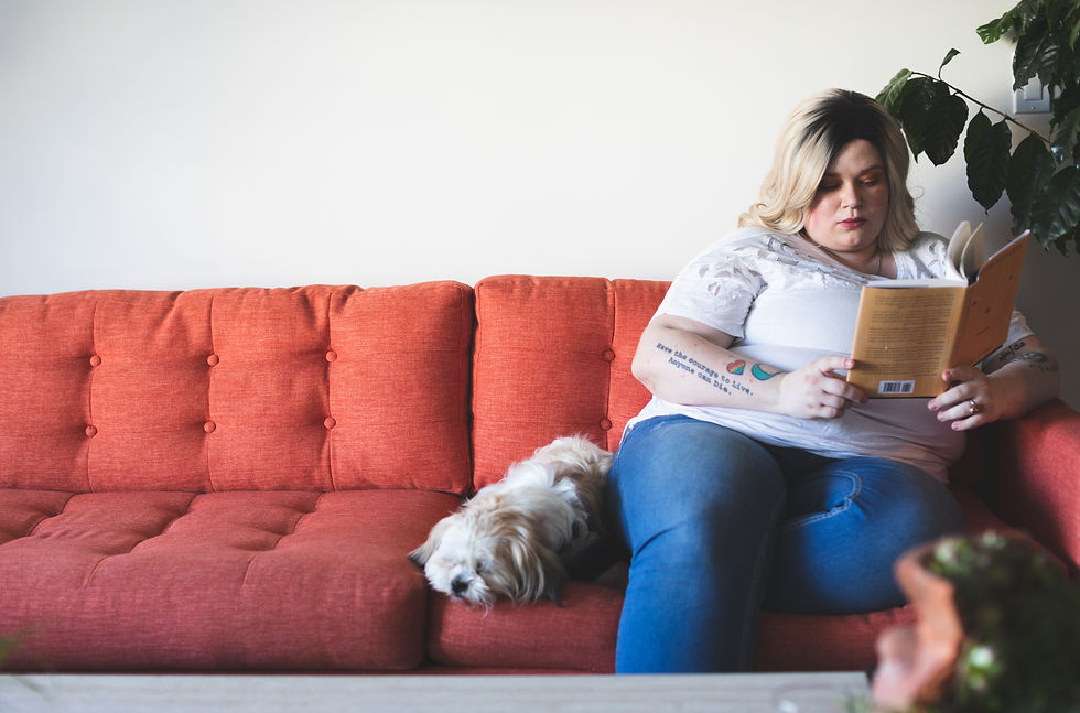 A larger bodied woman rests and reads on a couch next to a pet dog.