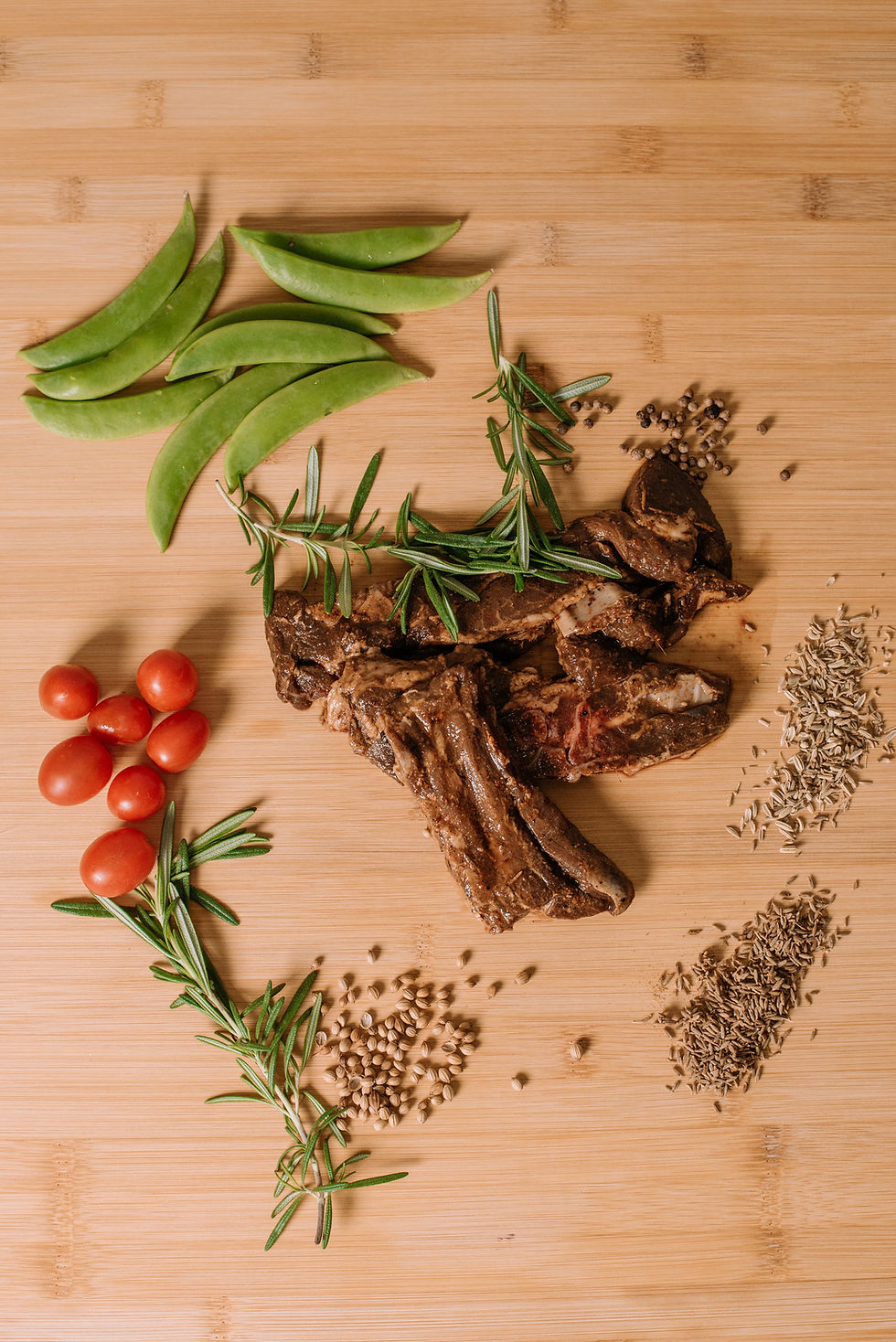 Grilled ribs with rosemary, cherry tomatoes, snow peas, and mixed spices arranged on a wooden surface. Rustic and appetizing setting.