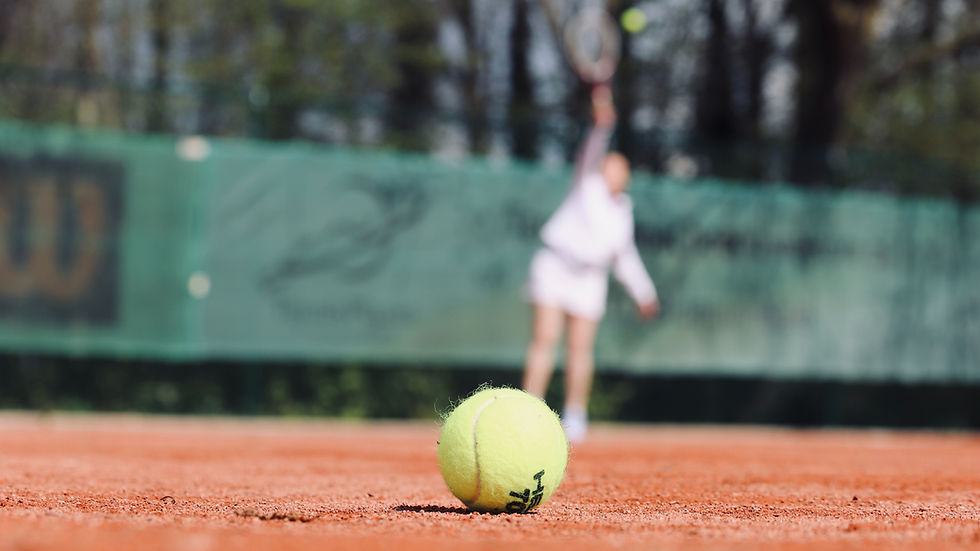 Eine junge Tennisspielerin unscharf im Hintergrund. Im Vordergrund ein Tennisball auf dem roten Sand des Tennisplatzes.