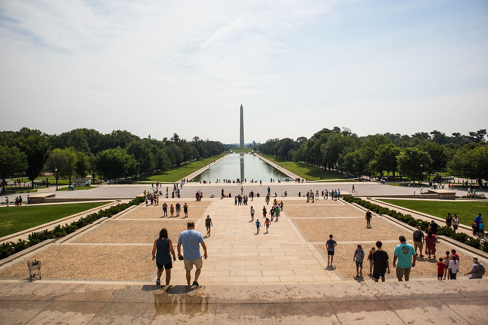 Tourists walk along the Lincoln Memorial Reflecting Pool toward the Washington Monument under a hazy sky.