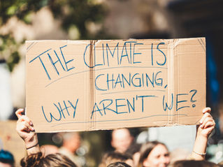The image shows a cardboard sign being held up with the words " The climate is changing, why aren't we?"