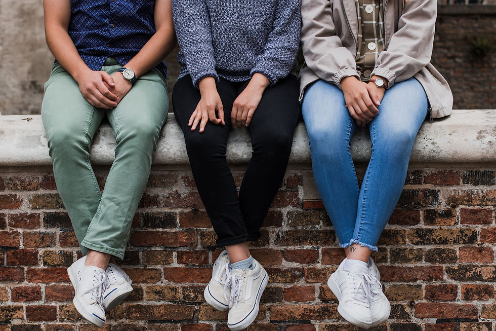 3 young people sitting on a brick wall wearing jeans.