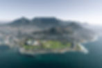 Luftaufnahme von Kapstadt: Tafelberg im Hintergrund, Stadt im Vordergrund, Stadion links, grüne Parks, blaues Meer. Klarer Himmel.