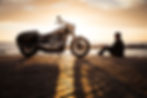 A man sits oceanside with his motorcycle at sunrise.