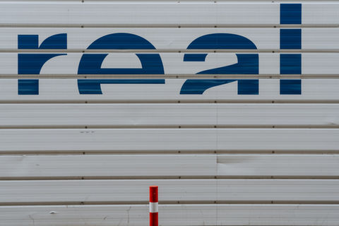 Large blue text "real" on a white corrugated wall, with a vertical red and white pole in the foreground. Industrial setting.