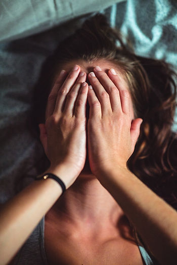 Person lying down with hands covering their face, conveying stress or fatigue, against a dark patterned background.